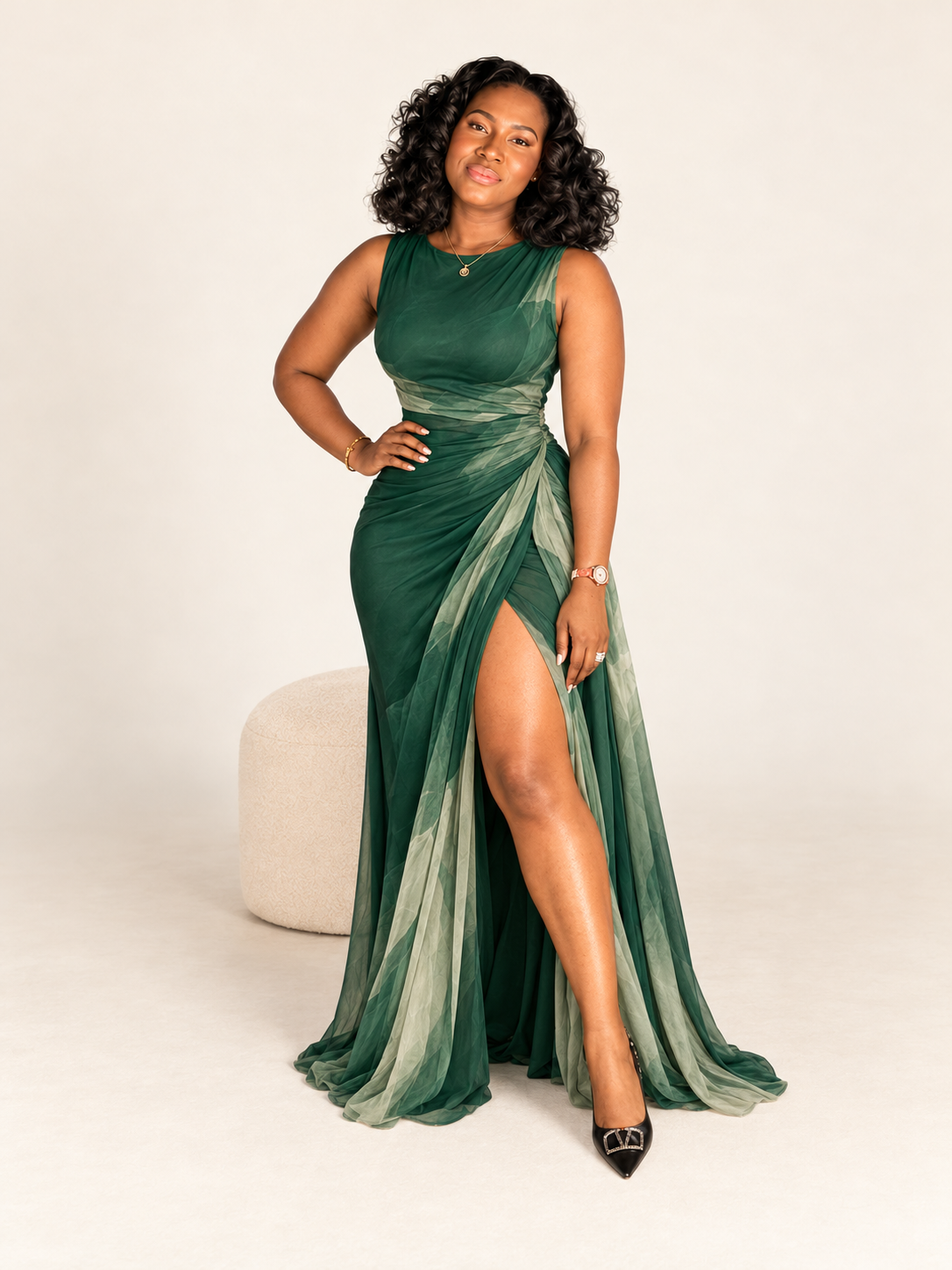 Portrait photograph of a woman in a floor-length emerald gown with sage draped overlays, standing with one hand on her waist against a warm ivory studio backdrop.